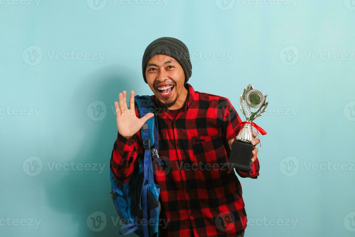 Excited Young Asian man student wearing a backpack, beanie hat, and red plaid flannel shirt, holding trophy, smiling at the camera, isolated on a blue background photo