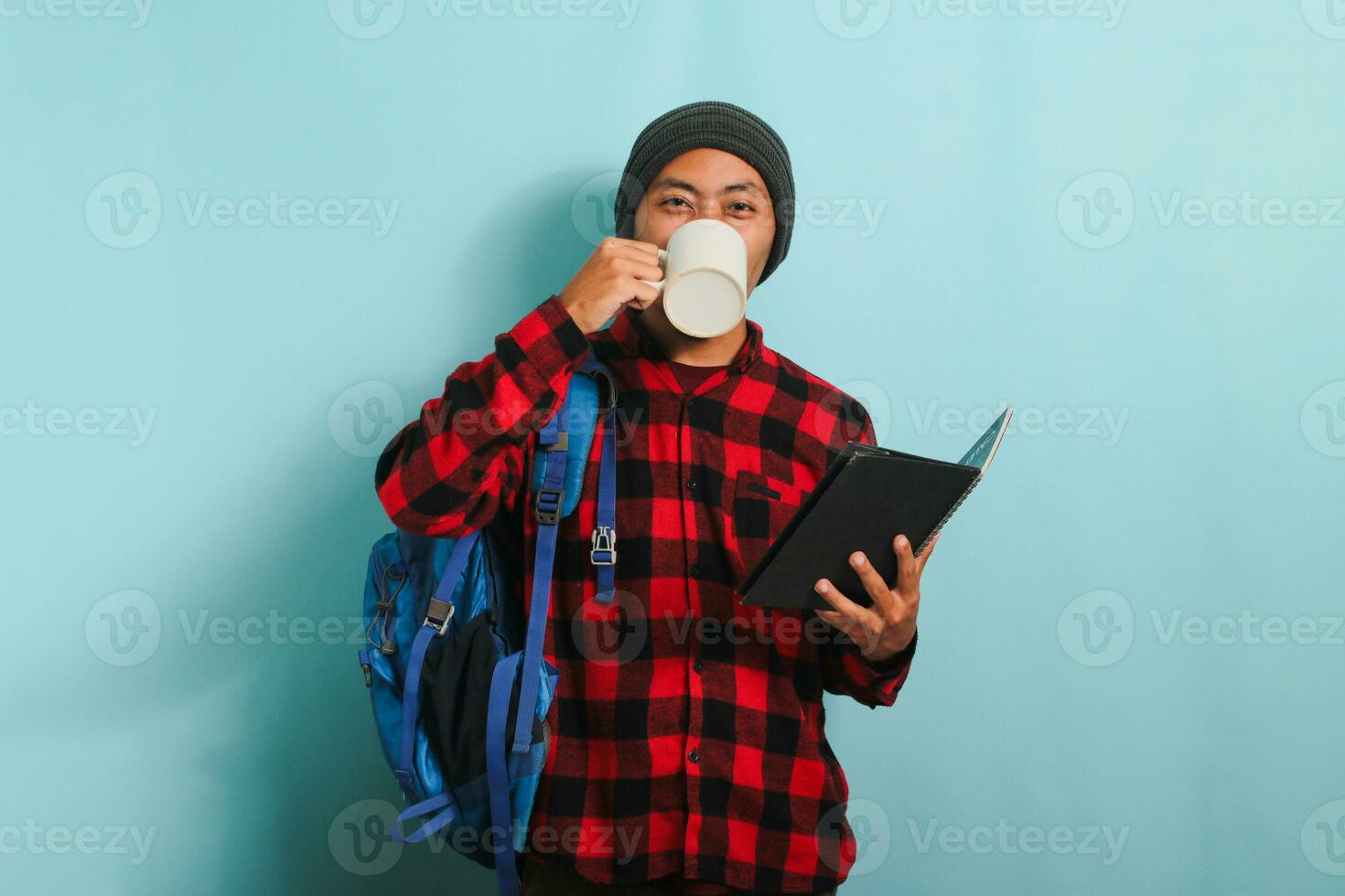 Happy young Asian male student with a beanie hat and a red plaid flannel shirt, wearing a backpack, takes a coffee break after lectures while standing against a blue background photo