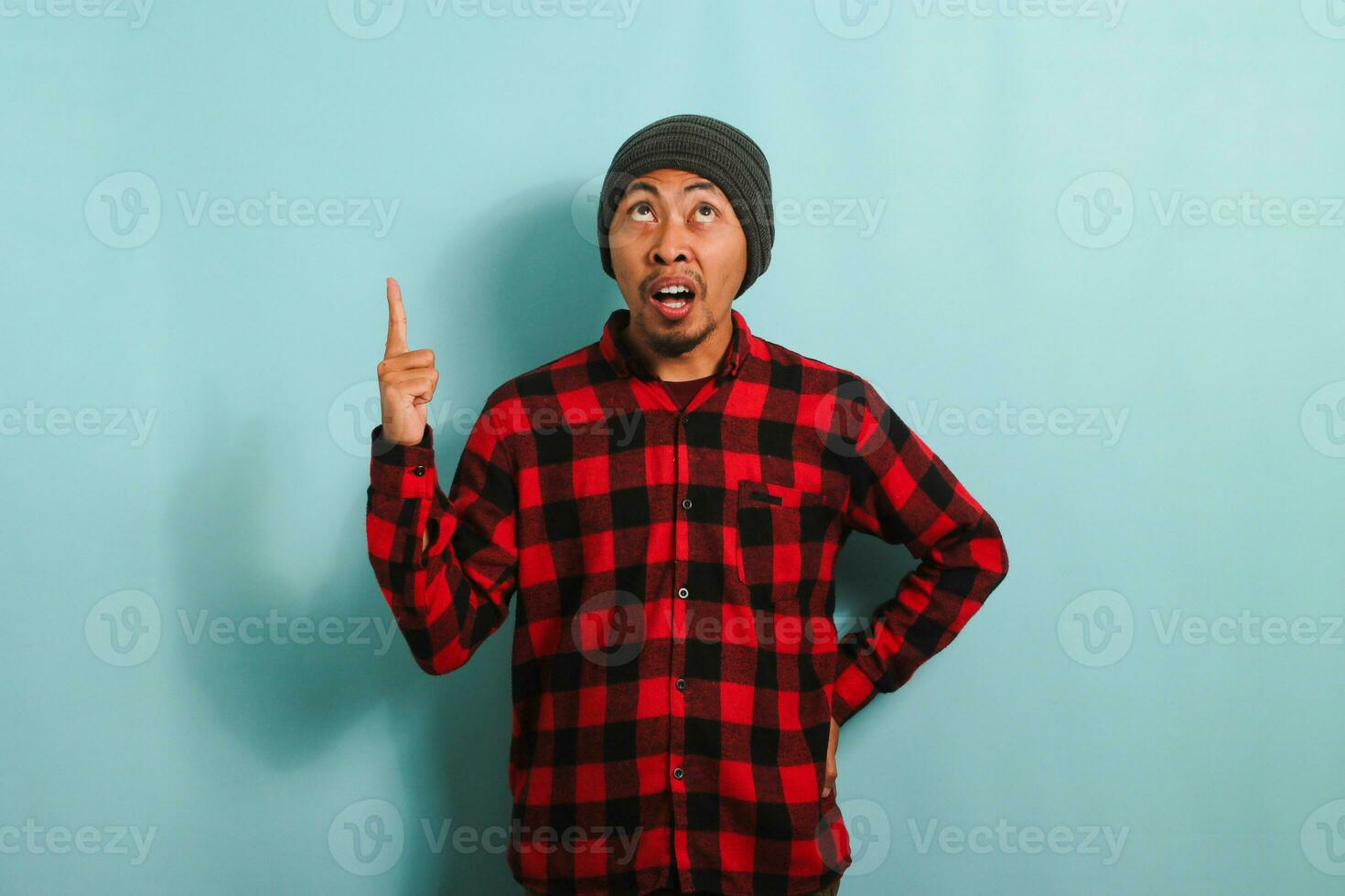 Surprised Young Asian man with a beanie hat and red plaid flannel shirt is pointing his finger up towards the copy space, isolated on a blue background photo