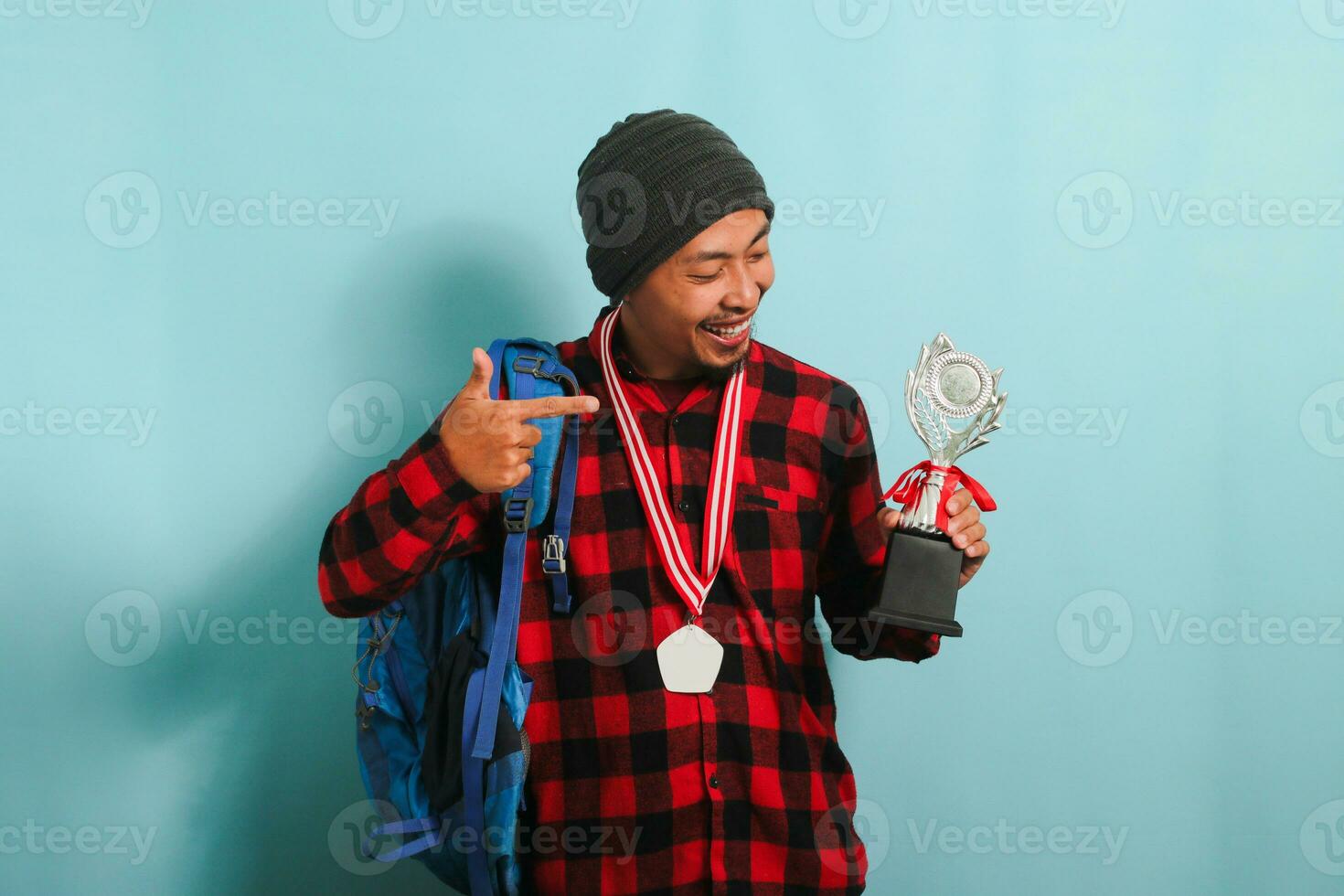 Excited young Asian man student wearing a backpack, beanie hat, and red plaid flannel shirt, pointing to the trophy in his hand while rejoicing in success and achievement, isolated on blue background photo