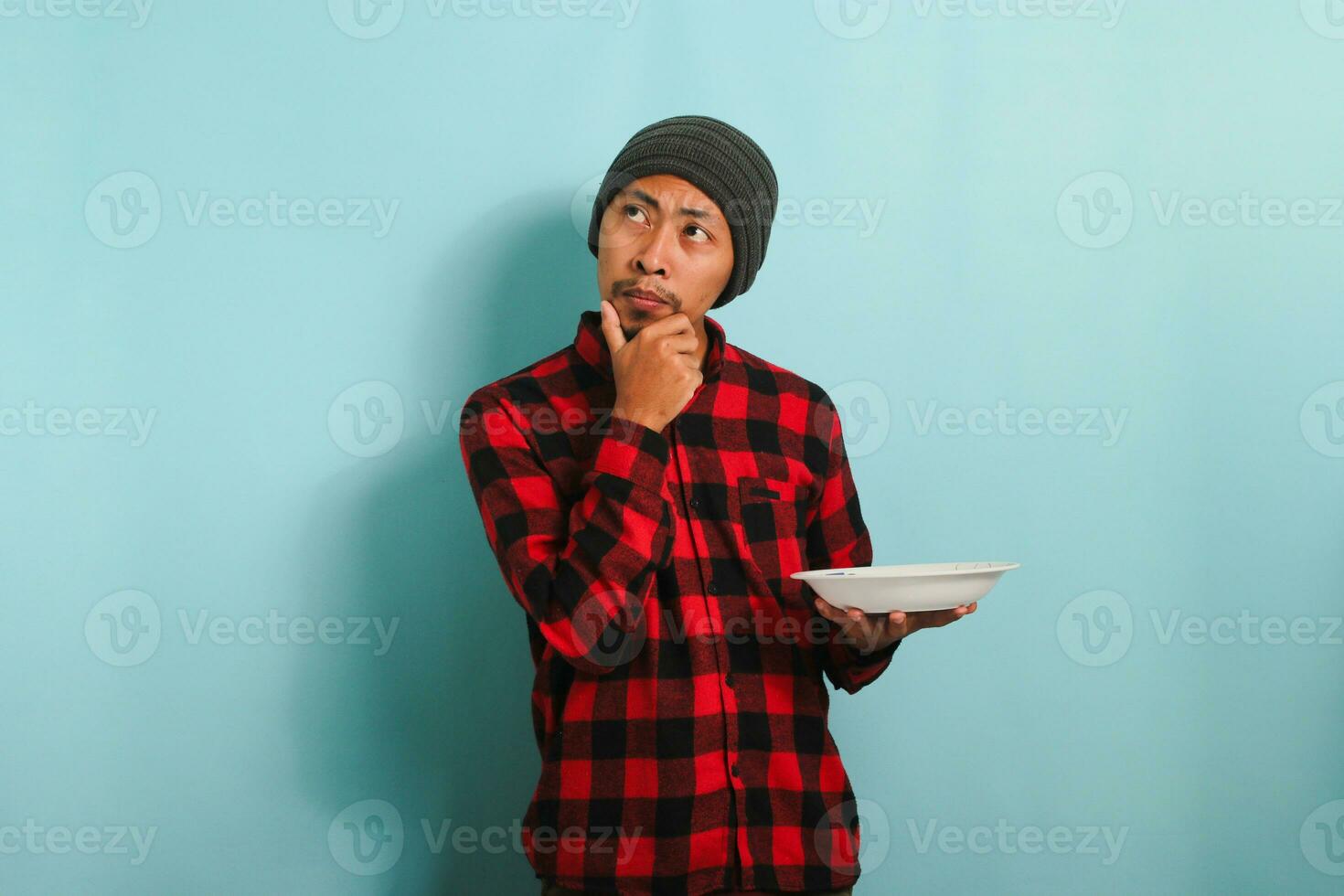 pensativo joven asiático hombre con gorro sombrero y rojo tartán franela camisa es sensación confundido, mirando aparte, pensando acerca de qué a comer mientras participación un vacío blanco lámina, aislado en un azul antecedentes foto