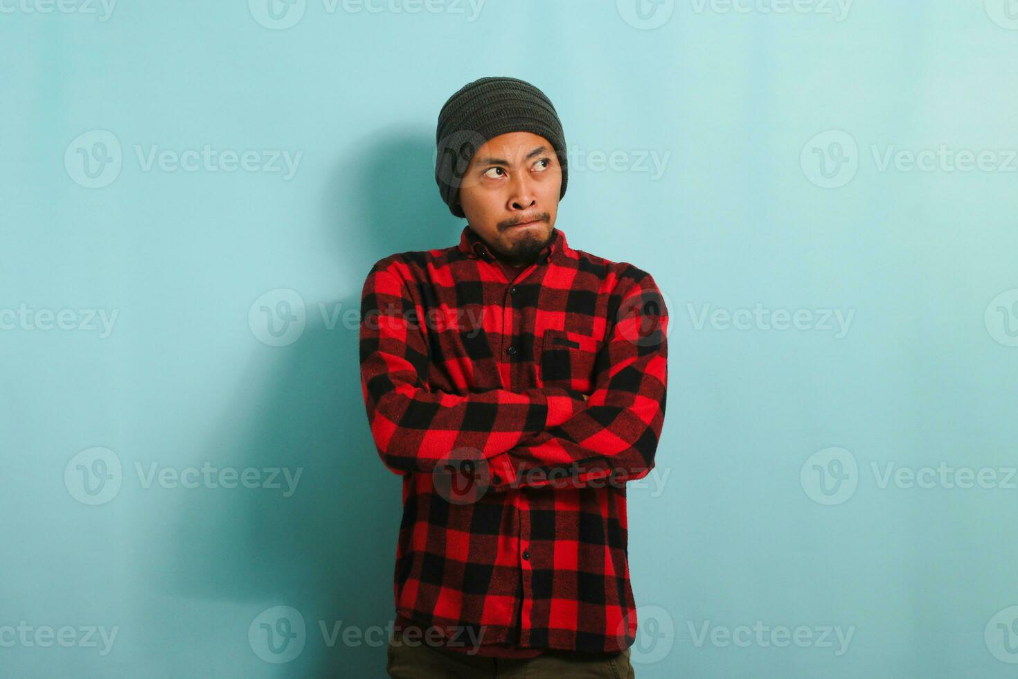 An angry young Asian man with a beanie hat and a red plaid flannel shirt keeps his arms folded, waiting for explanations with an annoyed and disapproving expression, isolated on a blue background photo