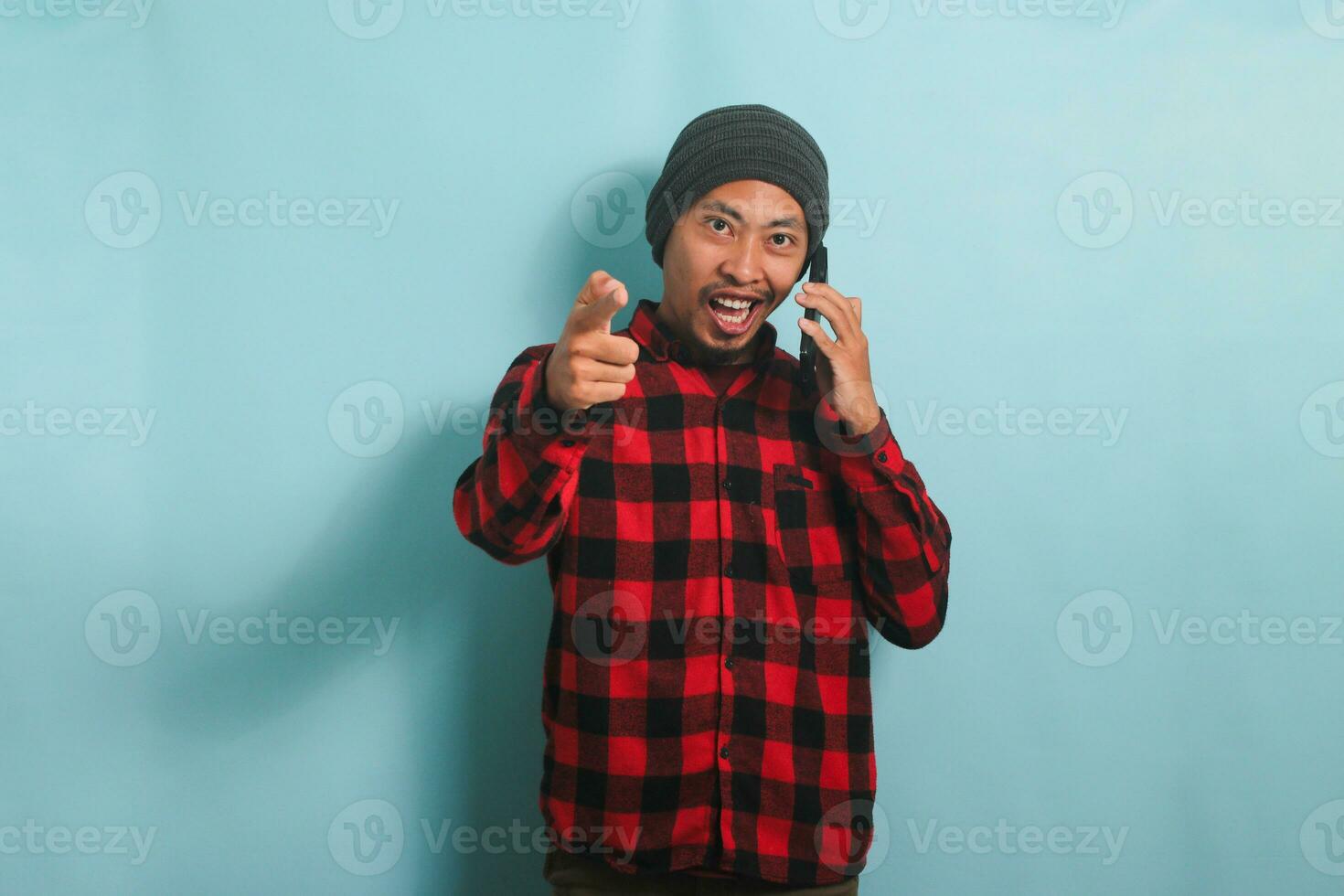Young Asian man with a beanie hat and a red plaid flannel shirt is smiling and pointing at the camera while talking on his mobile phone, isolated on a blue background photo