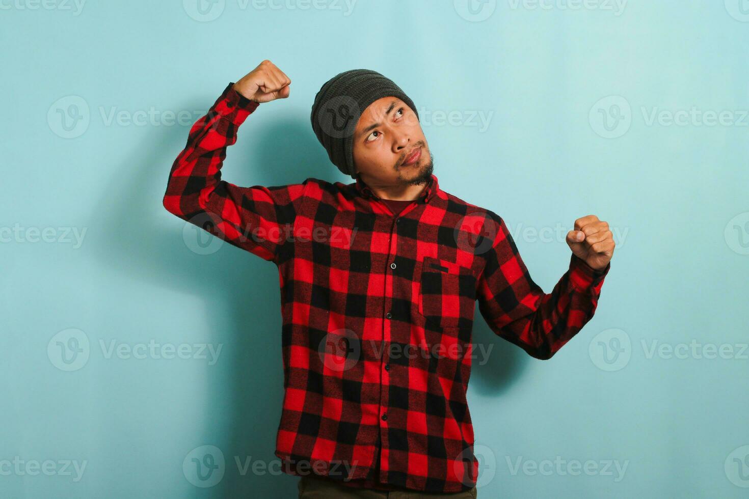 Young Asian man with beanie hat and red plaid flannel shirt making strong gesture, showing strength by flexing his arms and muscles, looking aside at empty copy space, isolated on a blue background photo