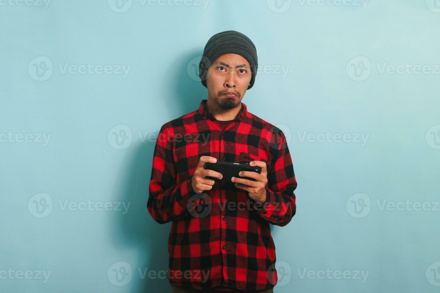 aburrido joven asiático hombre con un gorro sombrero y un rojo tartán franela camisa es jugando un aburrido juego y acecho un poco interesante vídeo en línea en su teléfono inteligente, aislado en un azul antecedentes foto