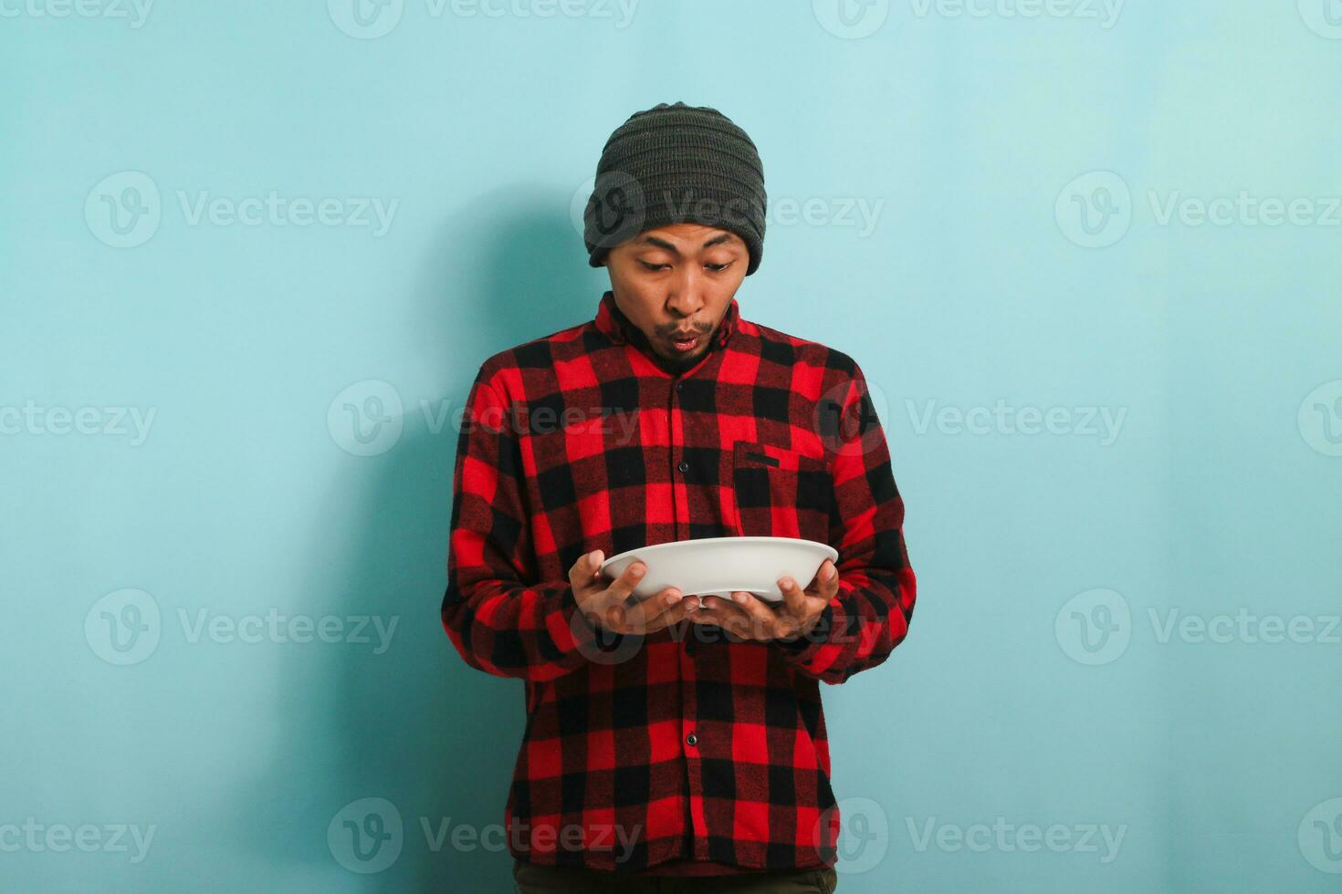 sorprendido joven asiático hombre con un gorro sombrero y un rojo tartán franela camisa es participación un vacío blanco plato en su mano, aislado en un azul antecedentes foto