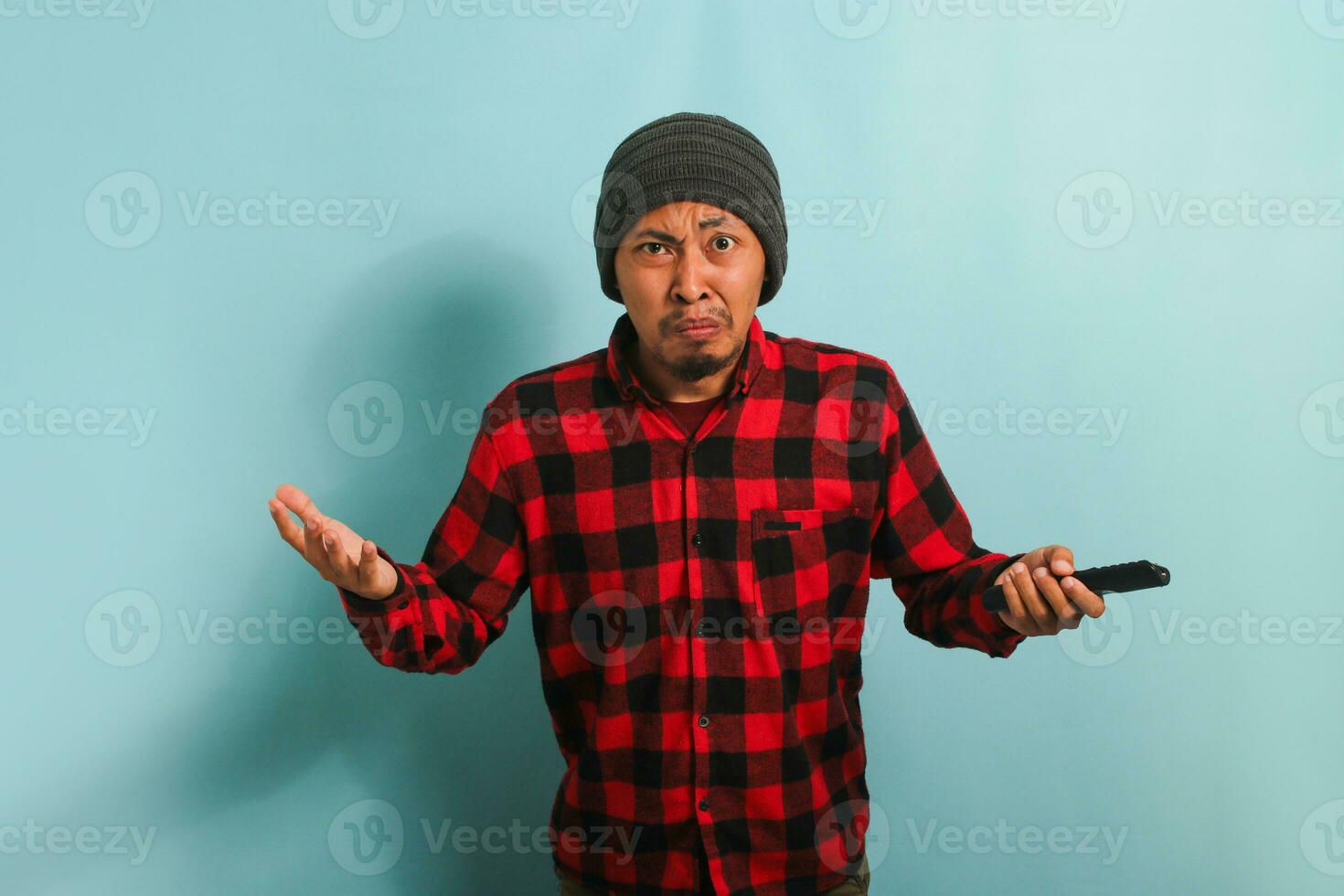 A furious young Asian man with a beanie hat and red plaid flannel shirt is arguing with the TV, getting angry while losing and watching a sports game on TV, isolated on a blue background photo