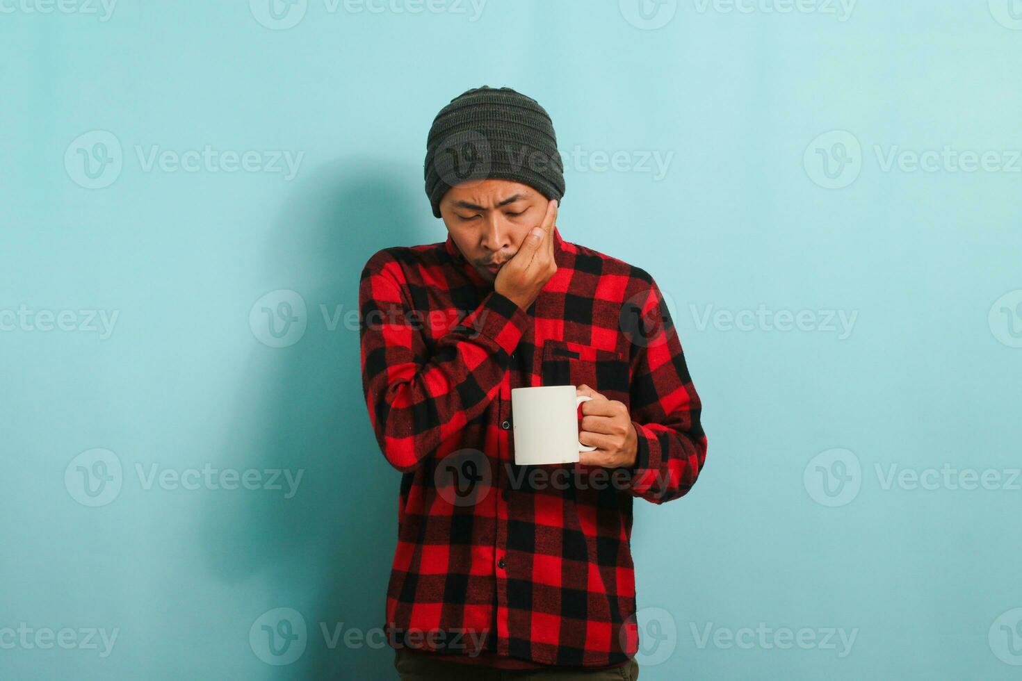 Unhappy Young Asian man with beanie hat and red plaid flannel shirt feeling terrible toothache after drinking cold water, suffering from sensitive toothache and cavities, isolated on blue background photo