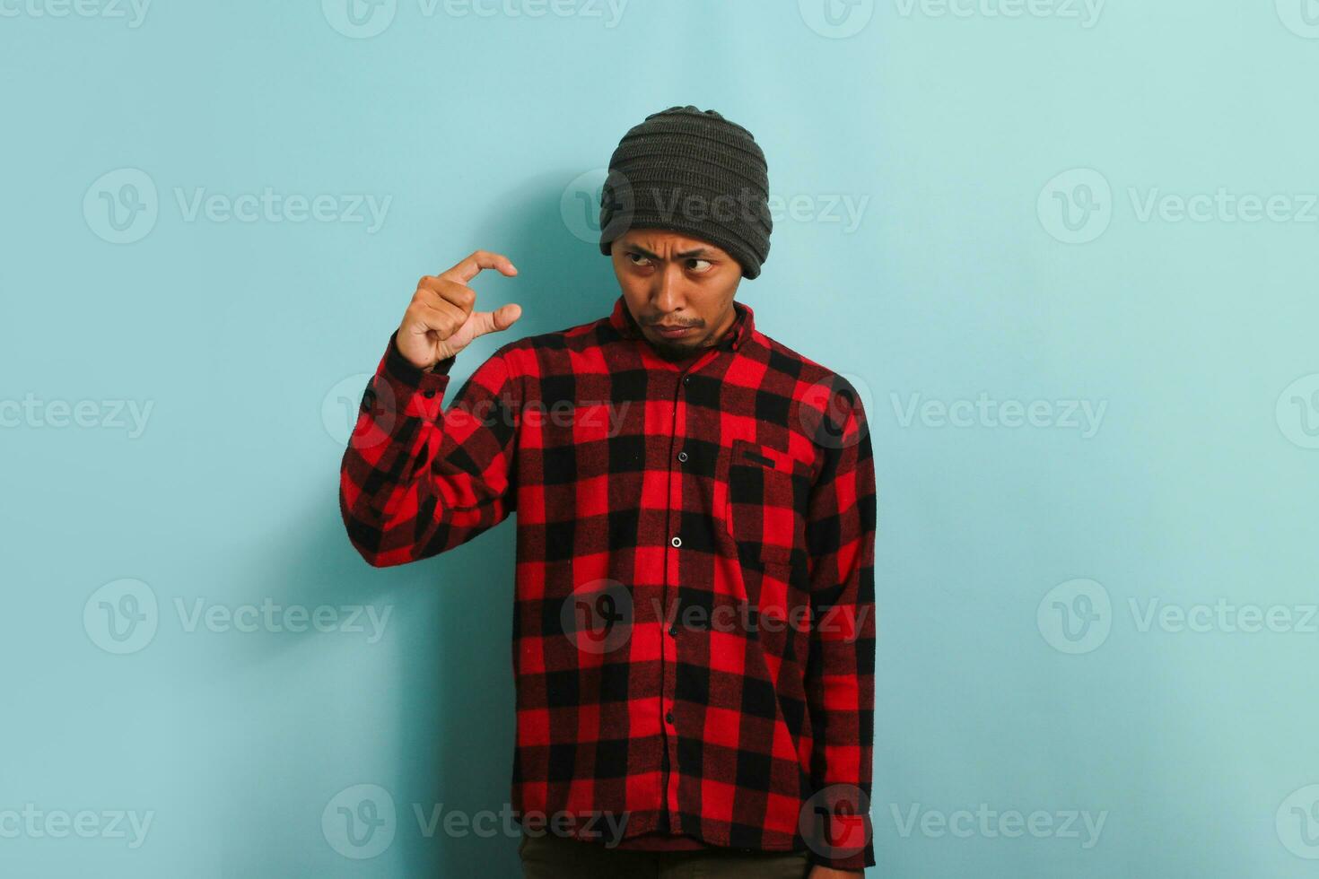 Confused Young Asian man with beanie hat and red plaid flannel shirt gesturing with his hand, showing small size sign with his fingers, representing a measurement symbol, isolated on a blue background photo