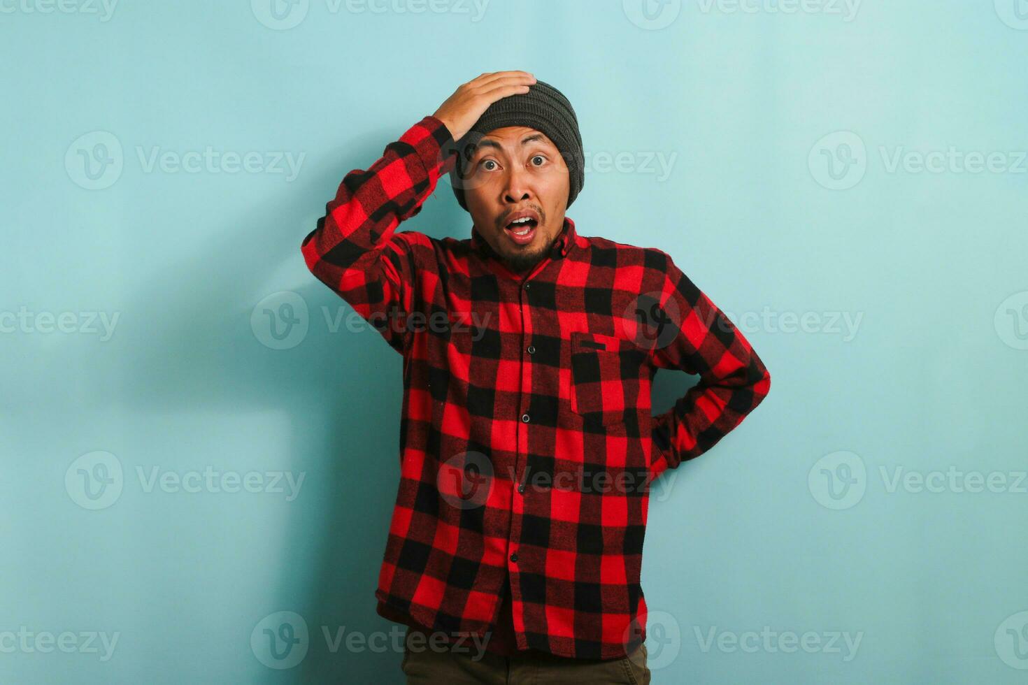 Scared Young Asian man with a beanie hat and red plaid flannel shirt has an open mouth, grabs his head, and looks at the camera reacting to something horrible, isolated on a blue background photo