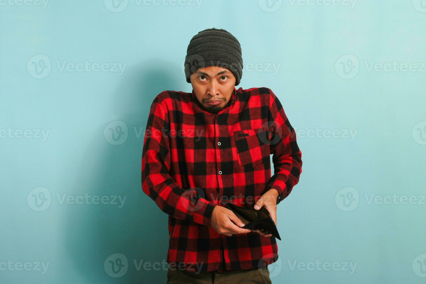 Shocked young Asian man with beanie hat and red plaid flannel shirt looking at his empty wallet with anger, isolated on a blue background photo