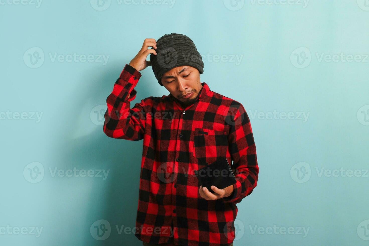 gracioso joven asiático hombre con gorro sombrero y rojo tartán franela camisa rascarse su cabeza en Confusión mientras mirando a su vacío billetera en choque, aislado en un azul antecedentes foto