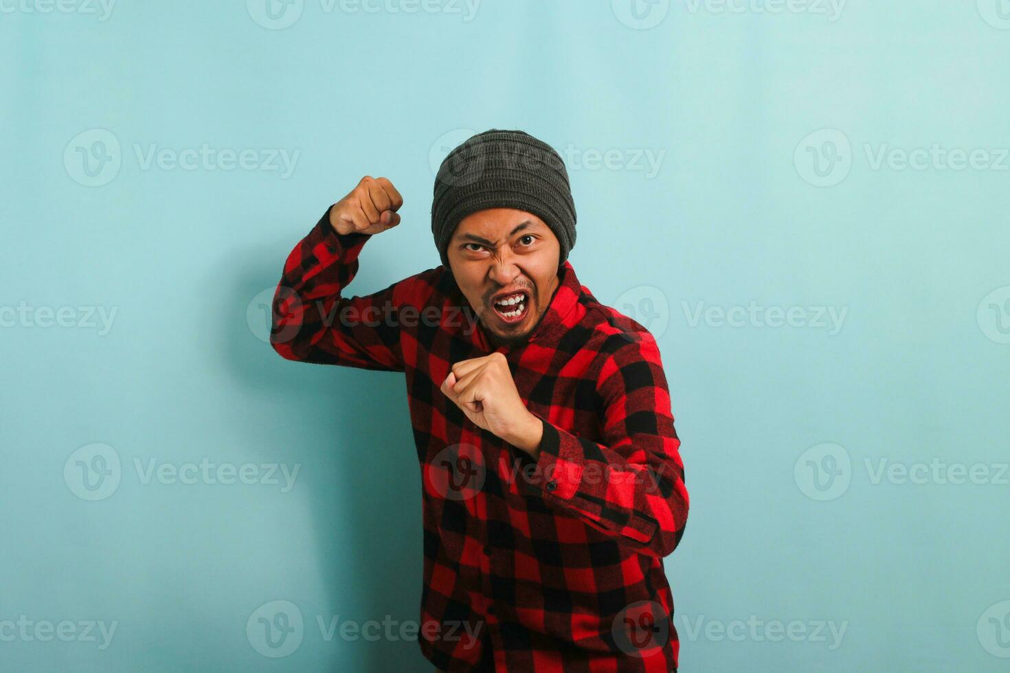 An angry, furious young Asian man with a beanie hat and a red plaid flannel shirt is ready to fight, with a defensive fist gesture, feeling mad and upset. He is isolated on a blue background photo