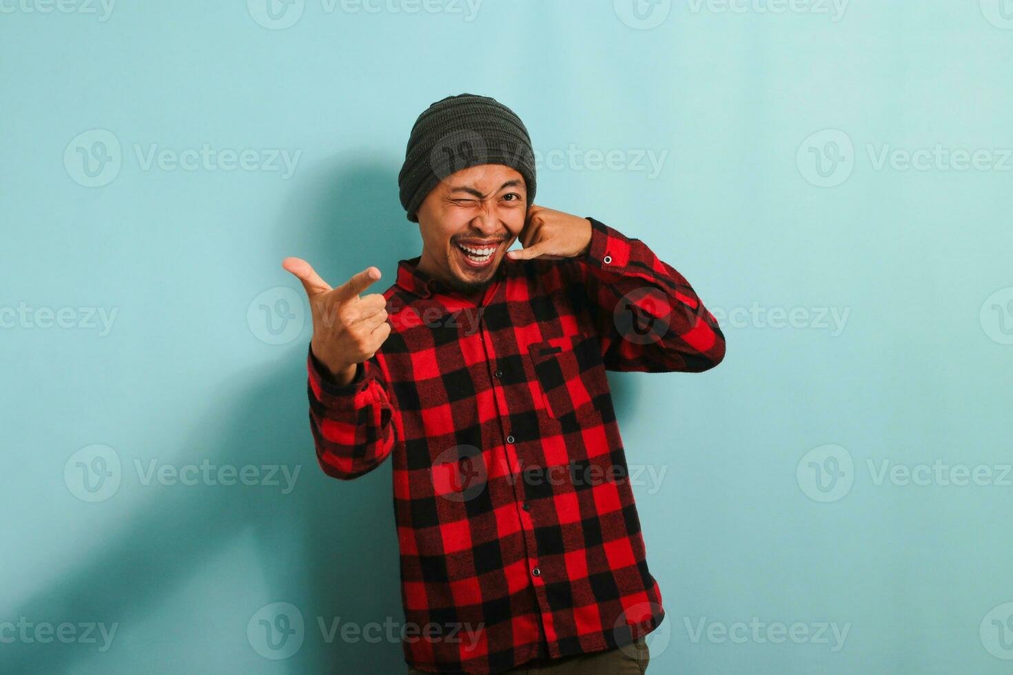 An excited young Asian man wearing a beanie hat and a red plaid flannel shirt makes a call me gesture while blinking and pointing at the camera, isolated on a blue background photo