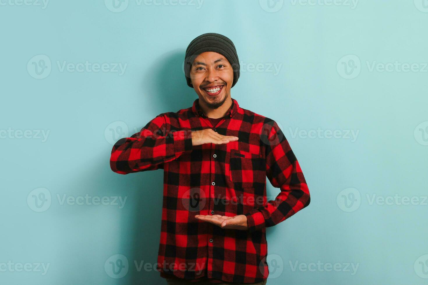 Young Asian man with beanie hat and red plaid flannel shirt gesturing a sign of size with both hands, measuring something, isolated on a blue background photo