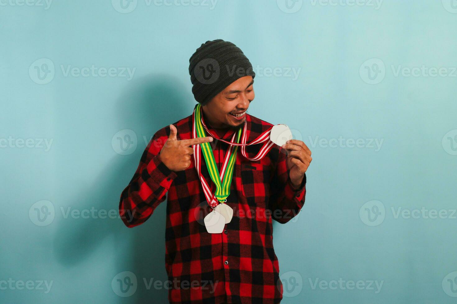 A young Asian man with a medal, a beanie hat, and a red plaid flannel shirt points a finger to himself, smiling with happiness and pride while standing against a blue background photo