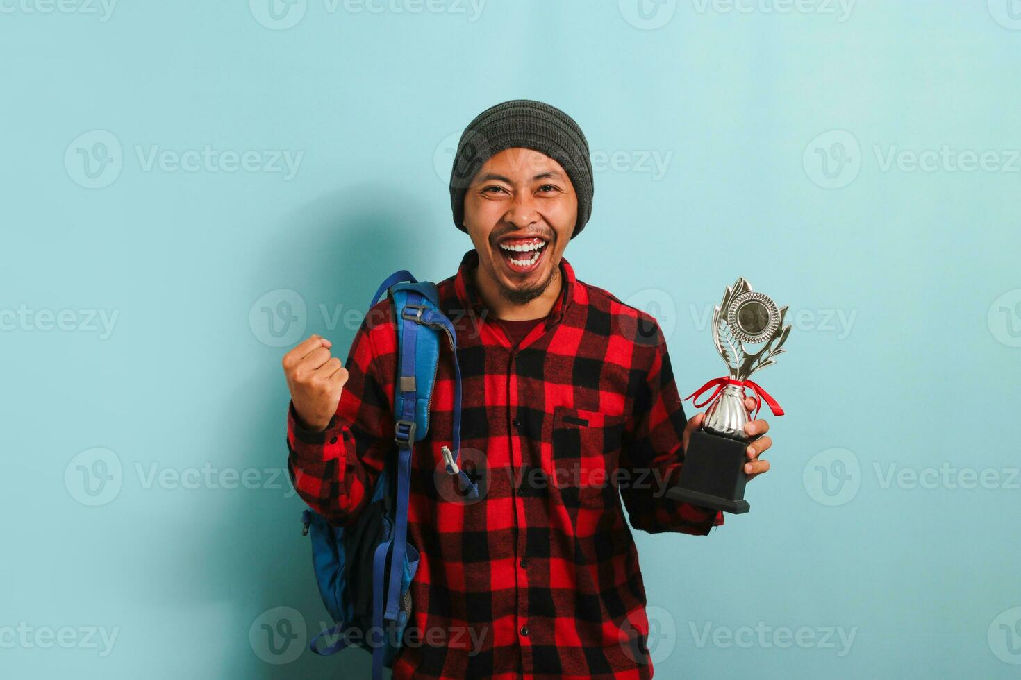 Excited young Asian man student wearing a backpack, beanie hat, and red plaid flannel shirt, making a YES gesture while holding a trophy and rejoicing in achievement, isolated on a blue background photo