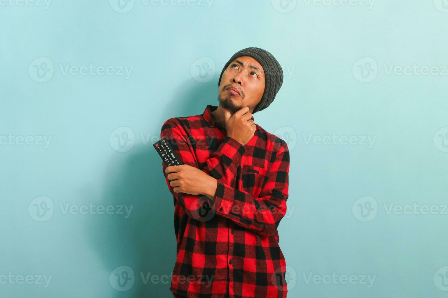 Pensive young Asian man with a beanie hat and red plaid flannel shirt thinking about what movie to watch while holding a TV remote, isolated on a blue background photo