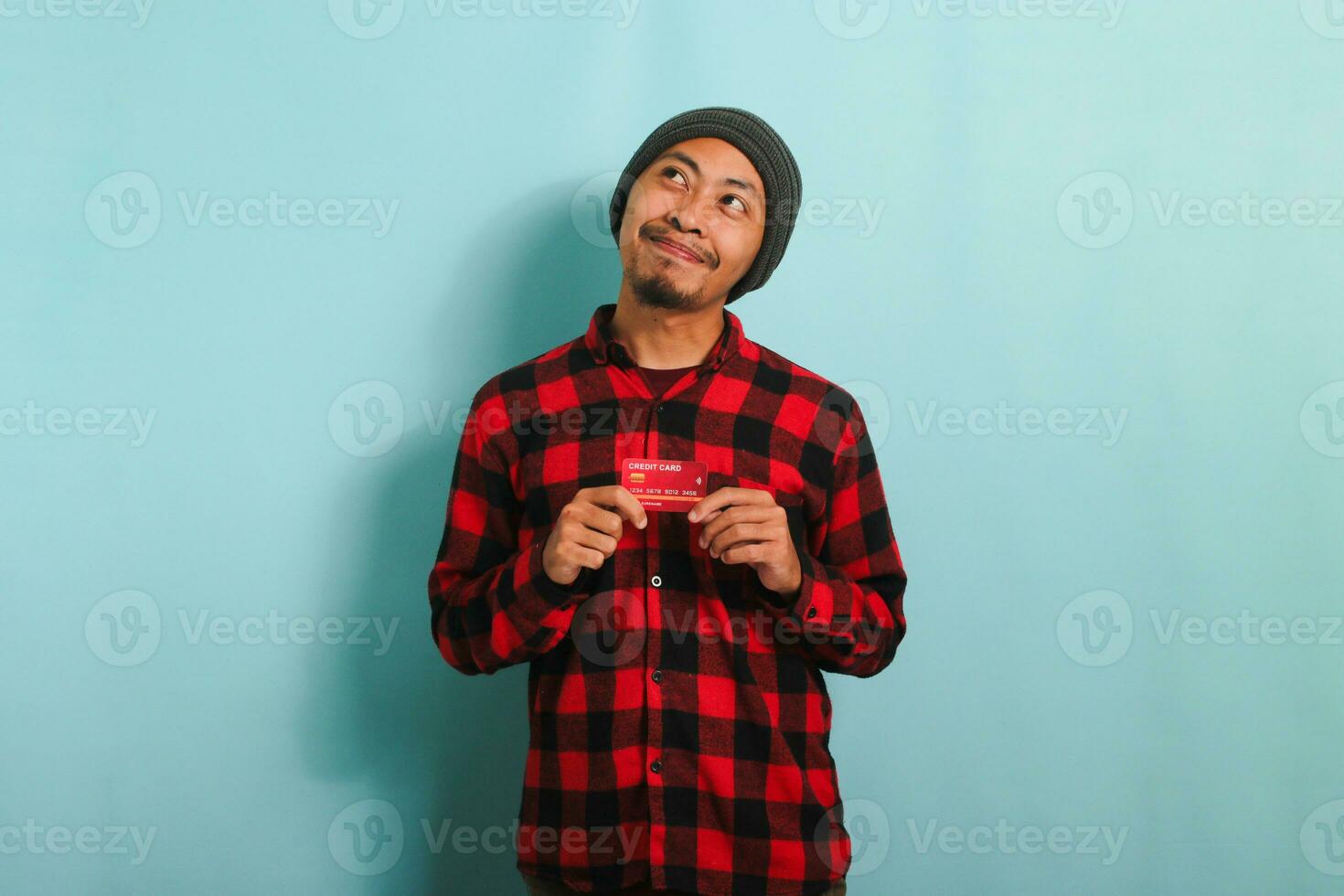 Pensive young Asian man with a beanie hat and red plaid flannel shirt is holding bank credit cards in his hands, looking at empty copy space, deep in thought, while standing against a blue background photo