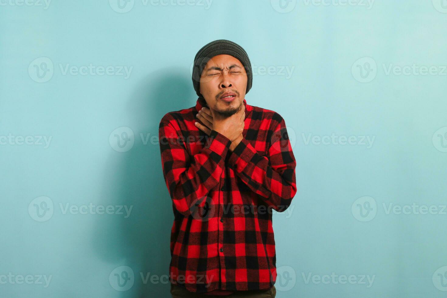 indispuesto joven asiático hombre con gorro sombrero y rojo tartán franela camisa participación su cuello, sufrimiento desde un dolorido garganta, frío, o gripe, aislado en un azul antecedentes foto