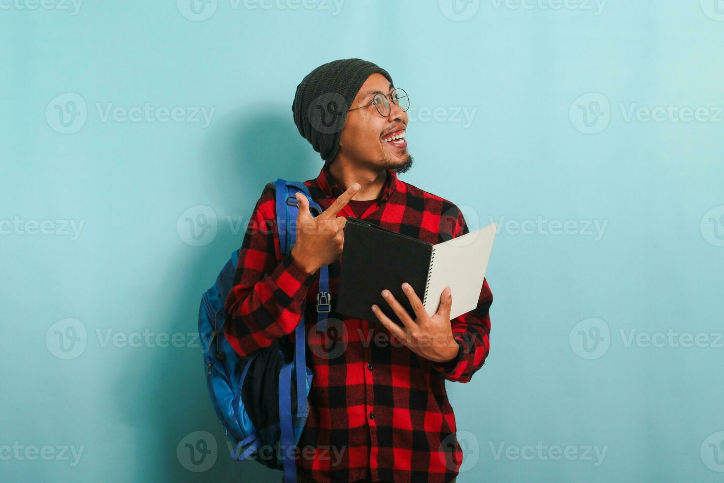 Excited young Asian student with a beanie hat and a red plaid flannel shirt, wearing a backpack and glasses, is holding books and pointing aside the copy space while standing against a blue background photo