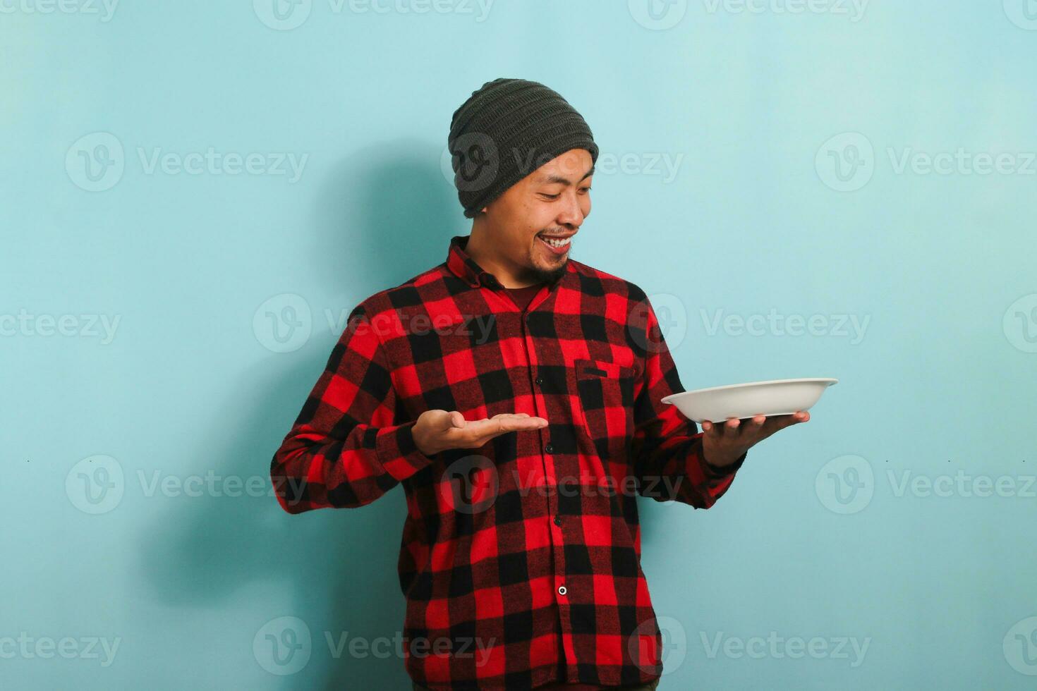 Excited Young Asian man with a beanie hat and a red plaid flannel shirt is showing an empty white plate in his hand, isolated on a blue background photo