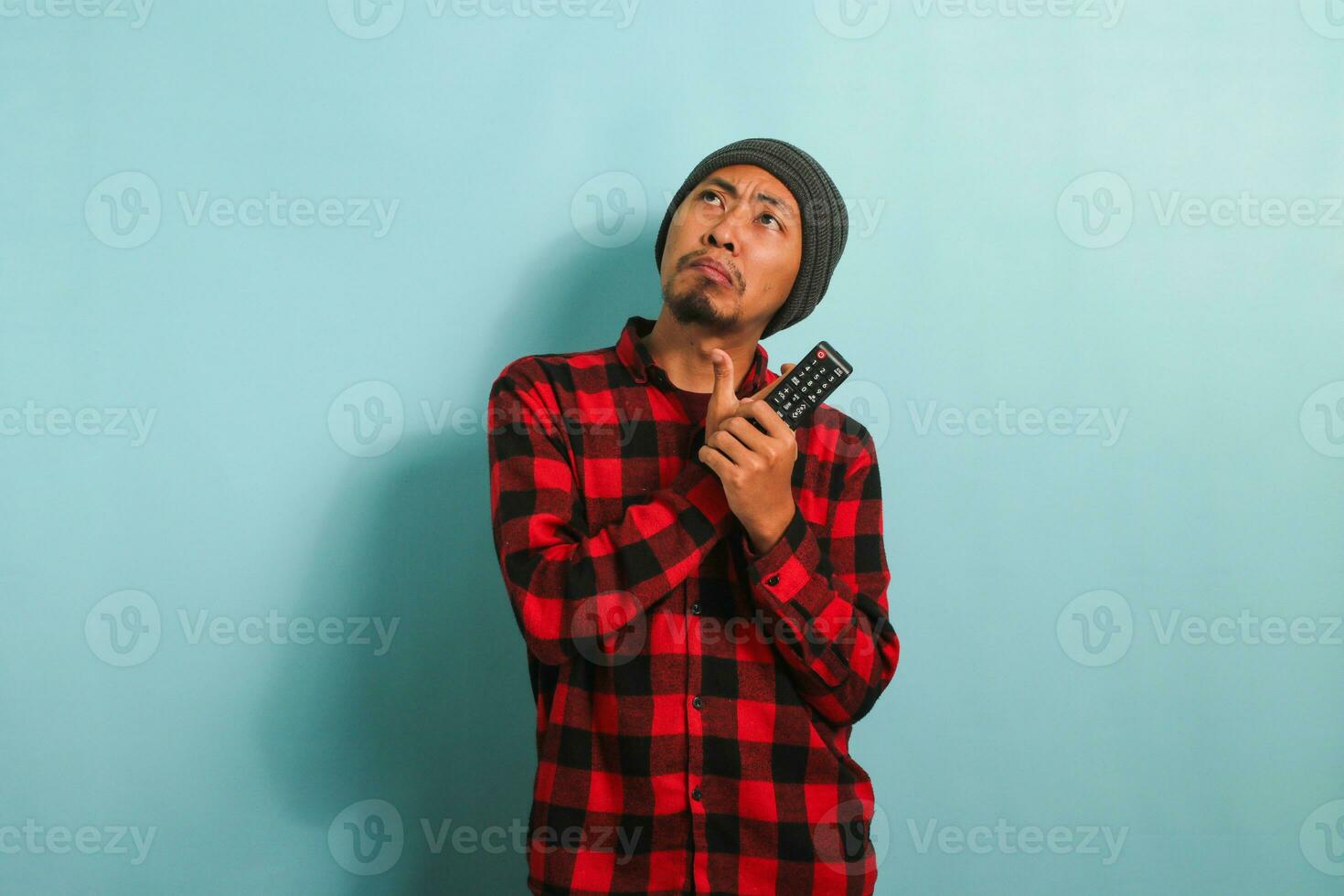 Pensive young Asian man with a beanie hat and red plaid flannel shirt thinking about what movie to watch while holding a TV remote, isolated on a blue background photo