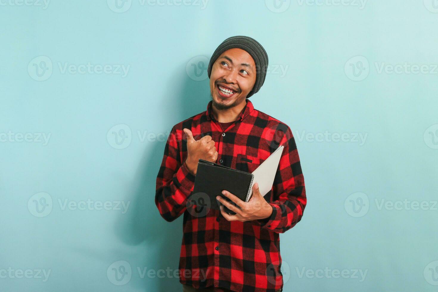 An excited young Asian man with a beanie hat and a red plaid flannel shirt is pointing his finger aside the copyspace while holding a book, standing against a blue background photo