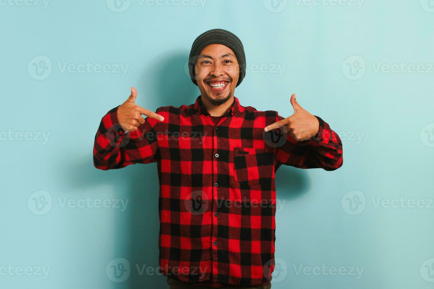 A confident young Asian man with a beanie hat and a red plaid flannel shirt is pointing at himself with his fingers, looking proud and happy. He is isolated on a blue background photo
