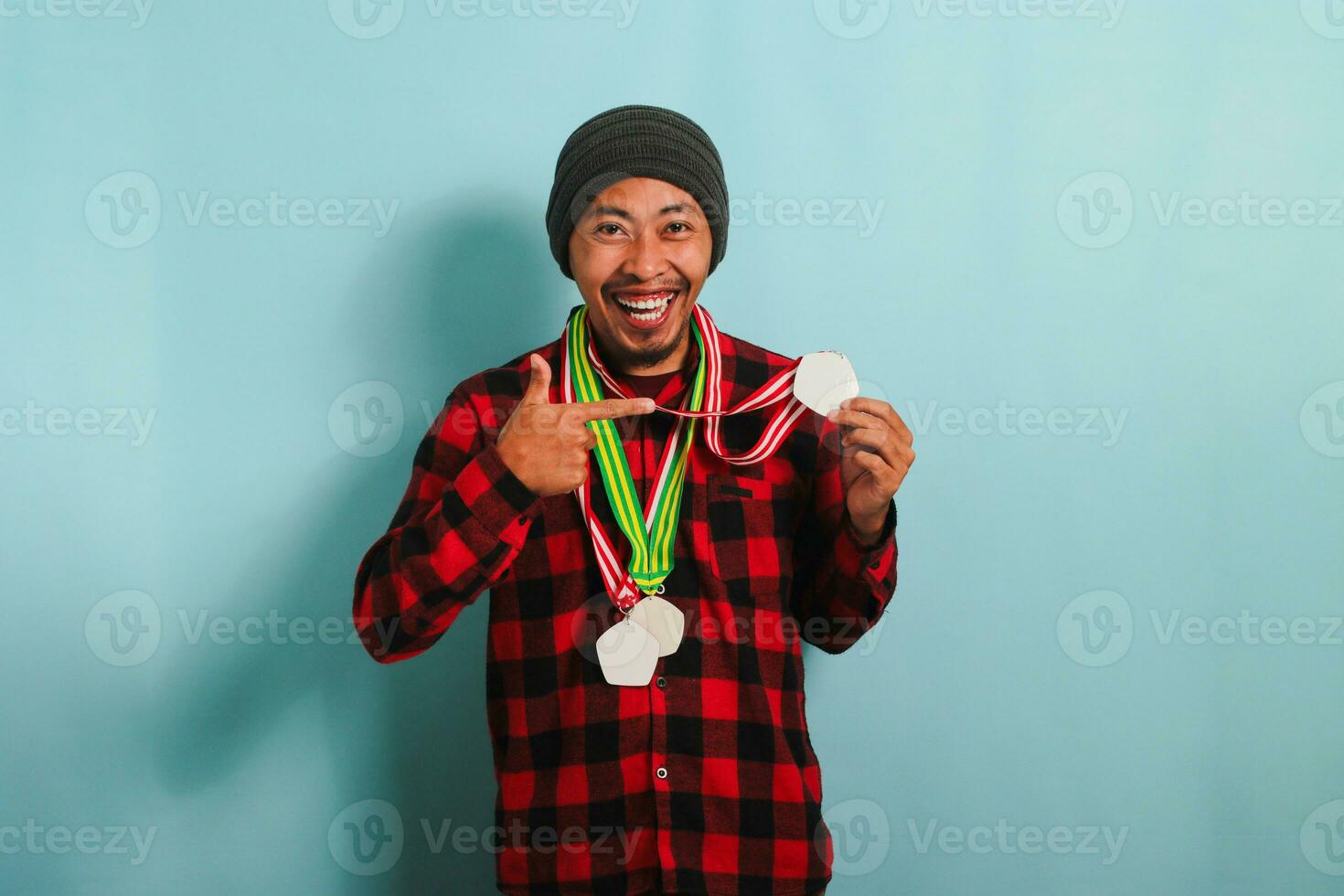 A young Asian man with a medal, a beanie hat, and a red plaid flannel shirt points a finger to himself, smiling with happiness and pride while standing against a blue background photo