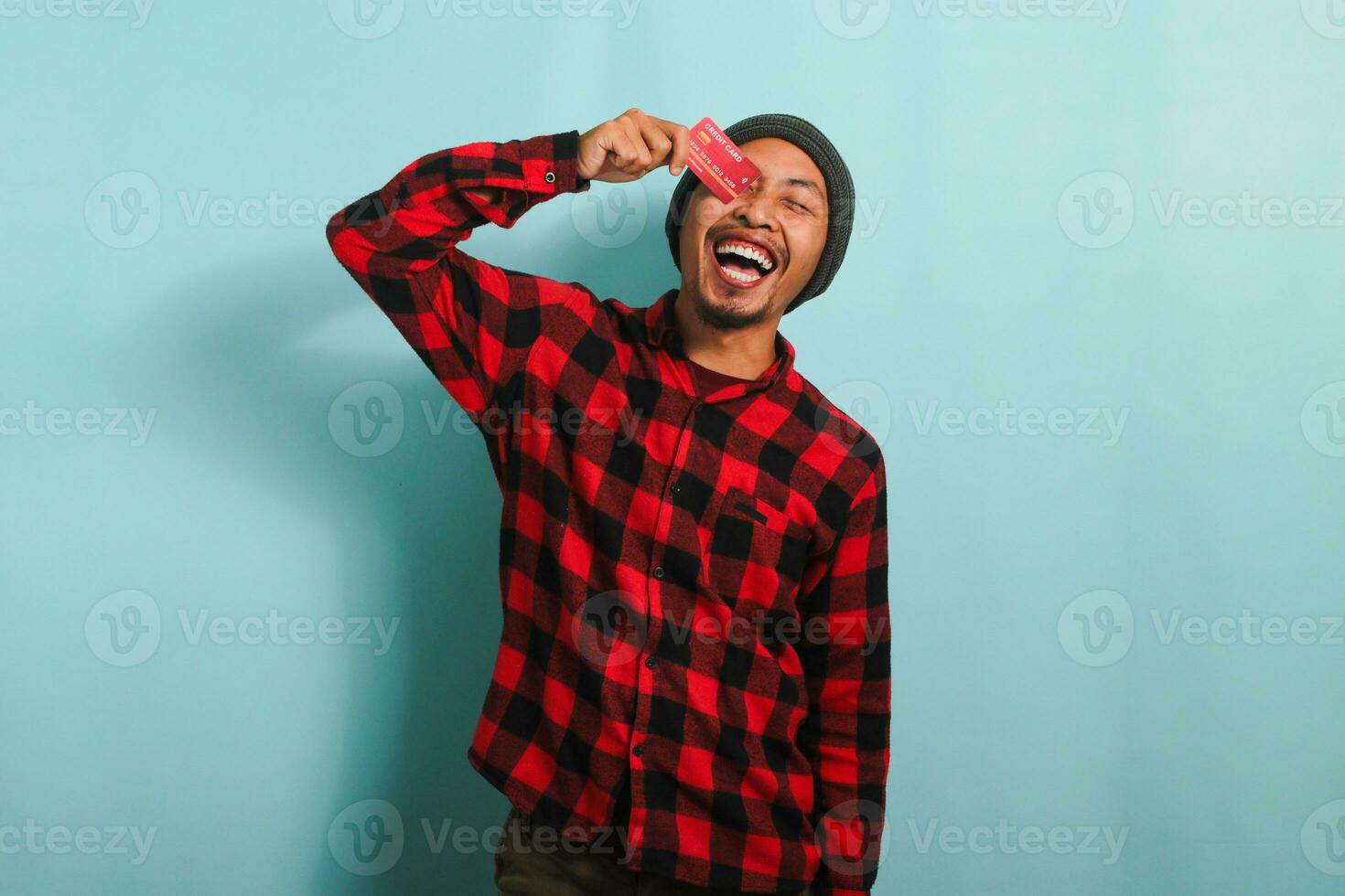 Joyful young Asian man with a beanie hat and red plaid flannel shirt is playfully covering one eye with a credit card while standing against a blue background photo