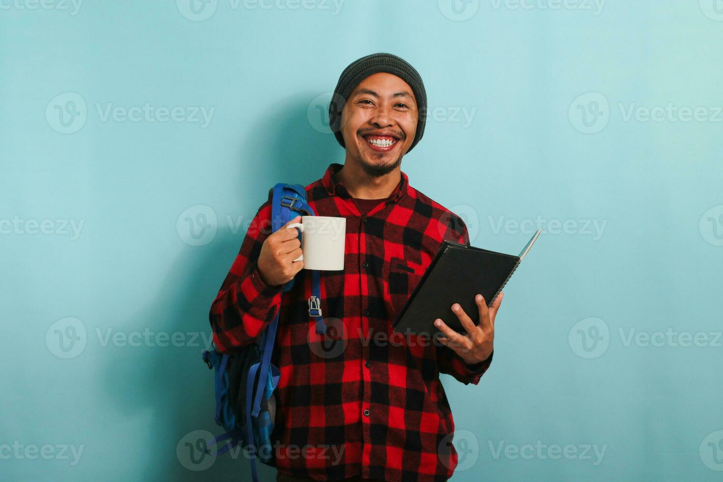 Happy young Asian male student with a beanie hat and a red plaid flannel shirt, wearing a backpack, takes a coffee break after lectures while standing against a blue background photo