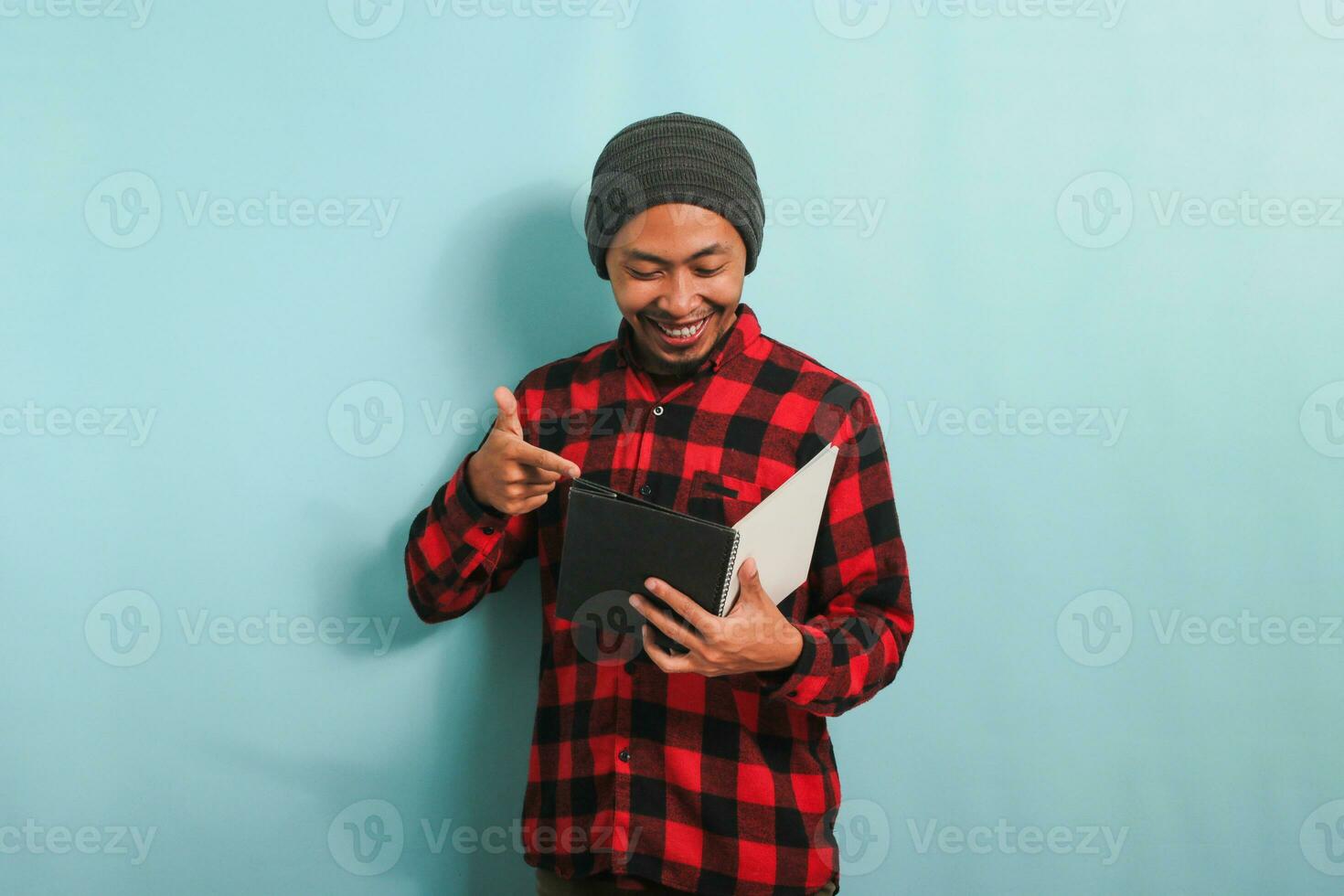 A smiling young Asian man with a beanie hat and a red plaid flannel shirt is pointing to a book in his hand while standing against a blue background photo