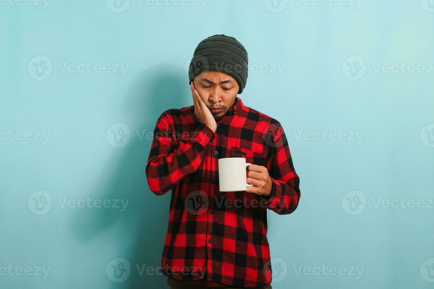 Unhappy Young Asian man with beanie hat and red plaid flannel shirt feeling terrible toothache after drinking cold water, suffering from sensitive toothache and cavities, isolated on blue background photo