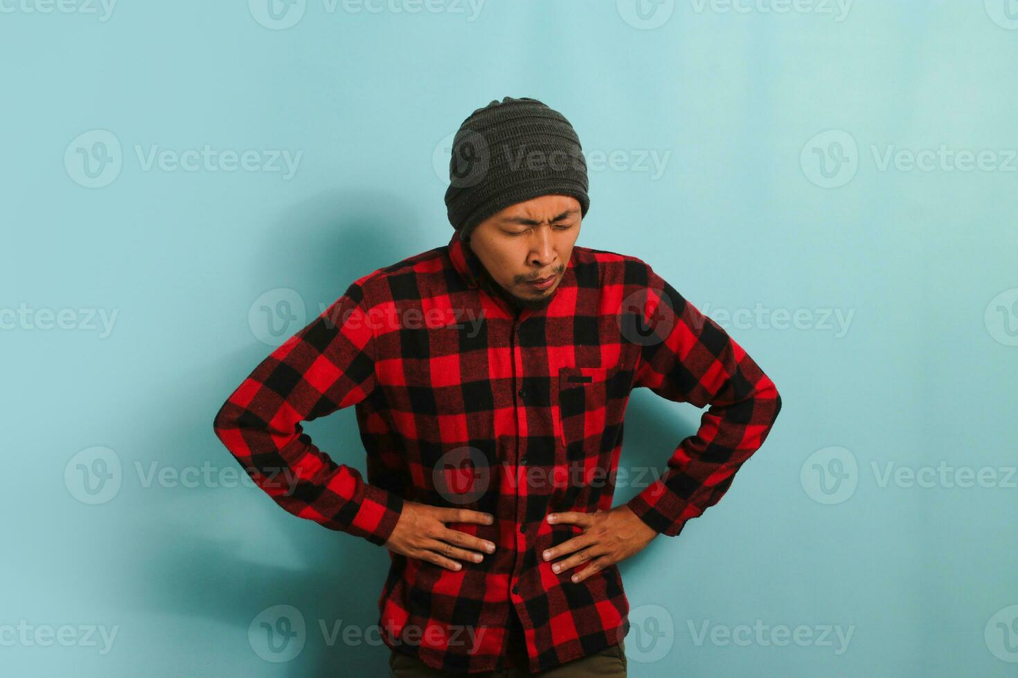 An unhappy young Asian man with a beanie hat and a red plaid flannel shirt is suffering from a stomach ache, bending over and holding his hands on his belly while standing against a blue background photo
