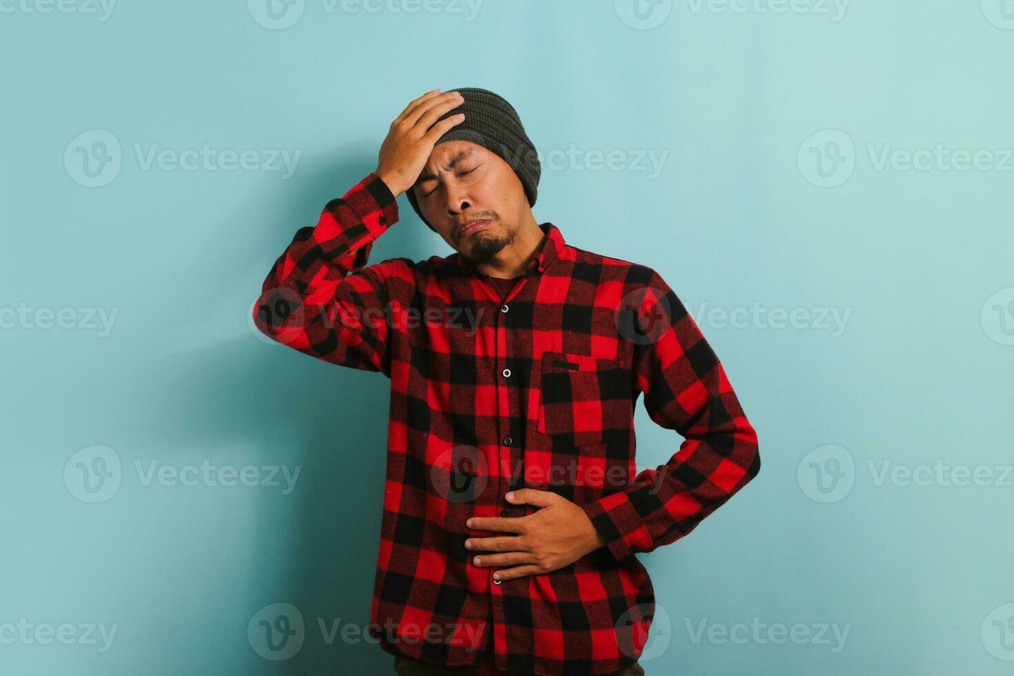 An unhappy young Asian man with a beanie hat and a red plaid flannel shirt is suffering from a stomach ache and feeling dizzy while standing against a blue background photo