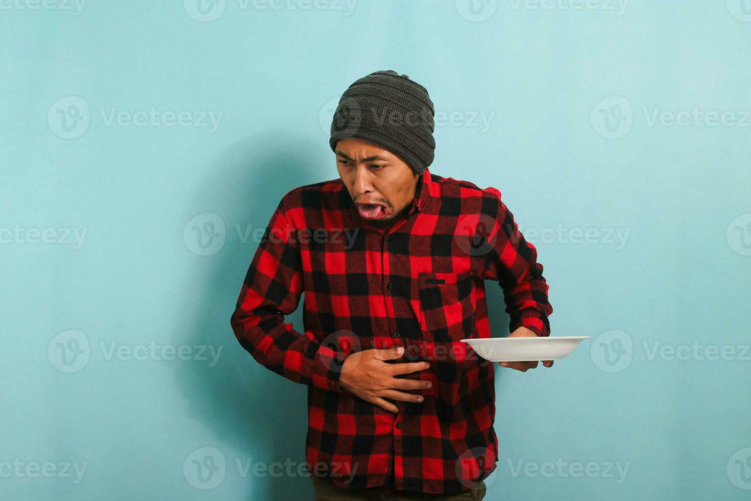 Young Asian man with a beanie hat and a red plaid flannel shirt is feeling nauseous while holding white plate, isolated on a blue background photo