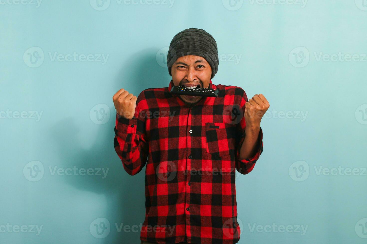 Funny young Asian man with a beanie hat and red plaid flannel shirt biting a TV remote, feeling angry while watching a sports game on TV, isolated on a blue background photo