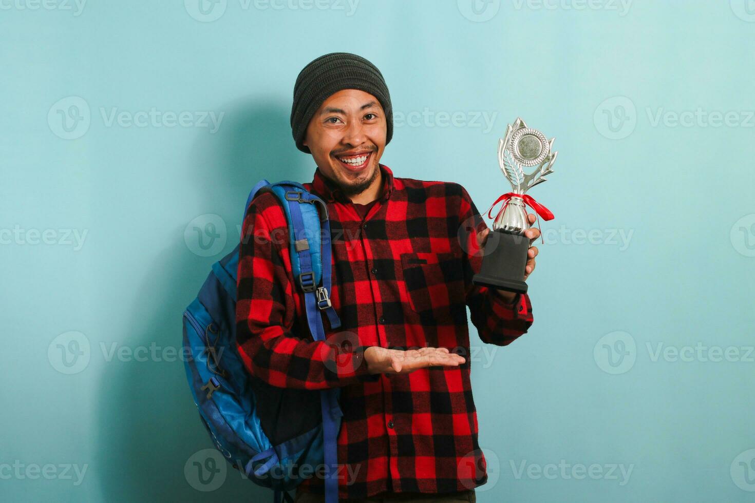Excited young Asian man student wearing a backpack, beanie hat, and red plaid flannel shirt, pointing to the trophy in his hand while rejoicing in success and achievement, isolated on blue background photo