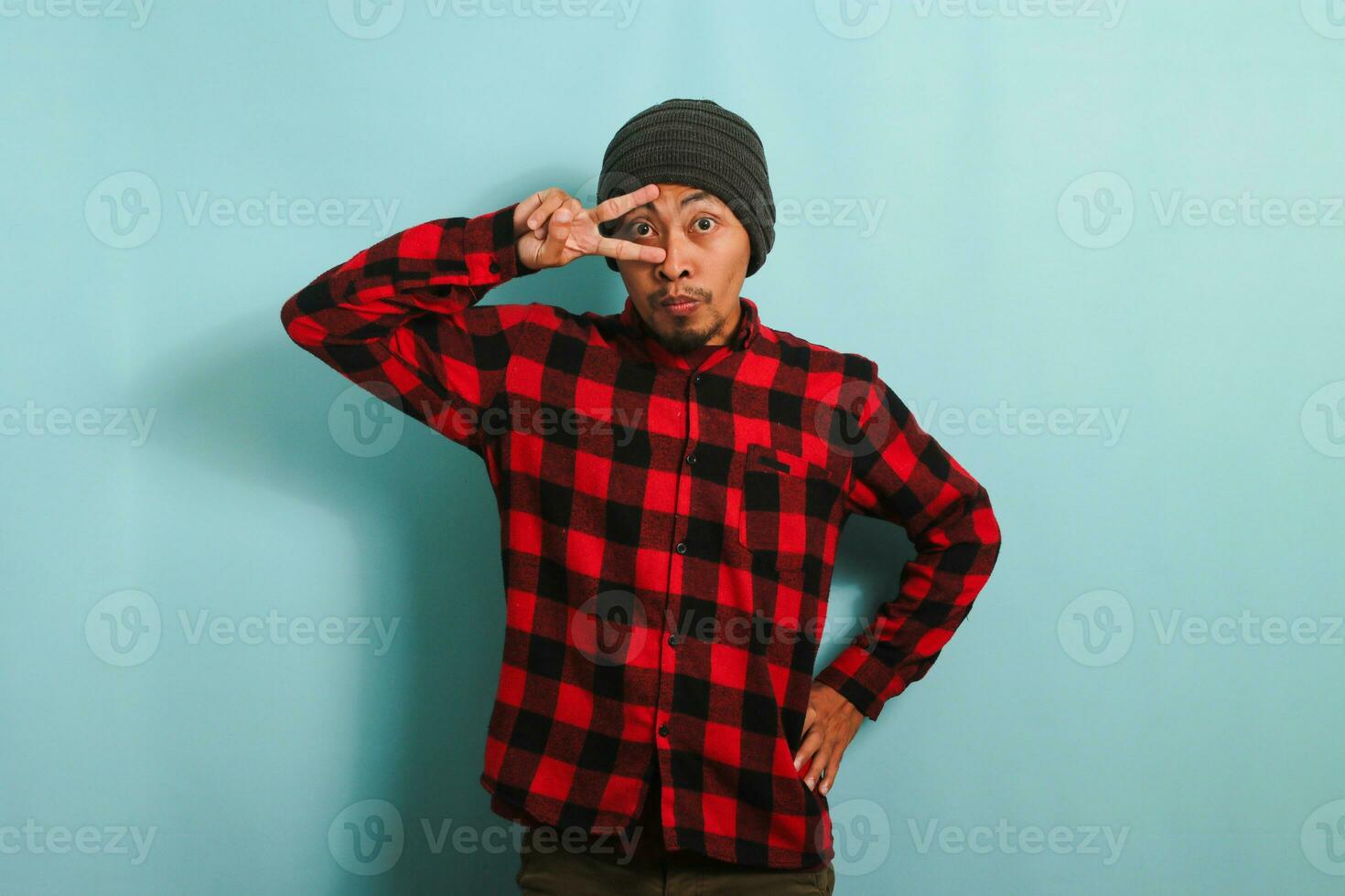 Attractive young Asian man with a beanie hat and a red plaid flannel shirt poses for the camera with a smile on his face and shows a peace gesture while standing against a blue background photo