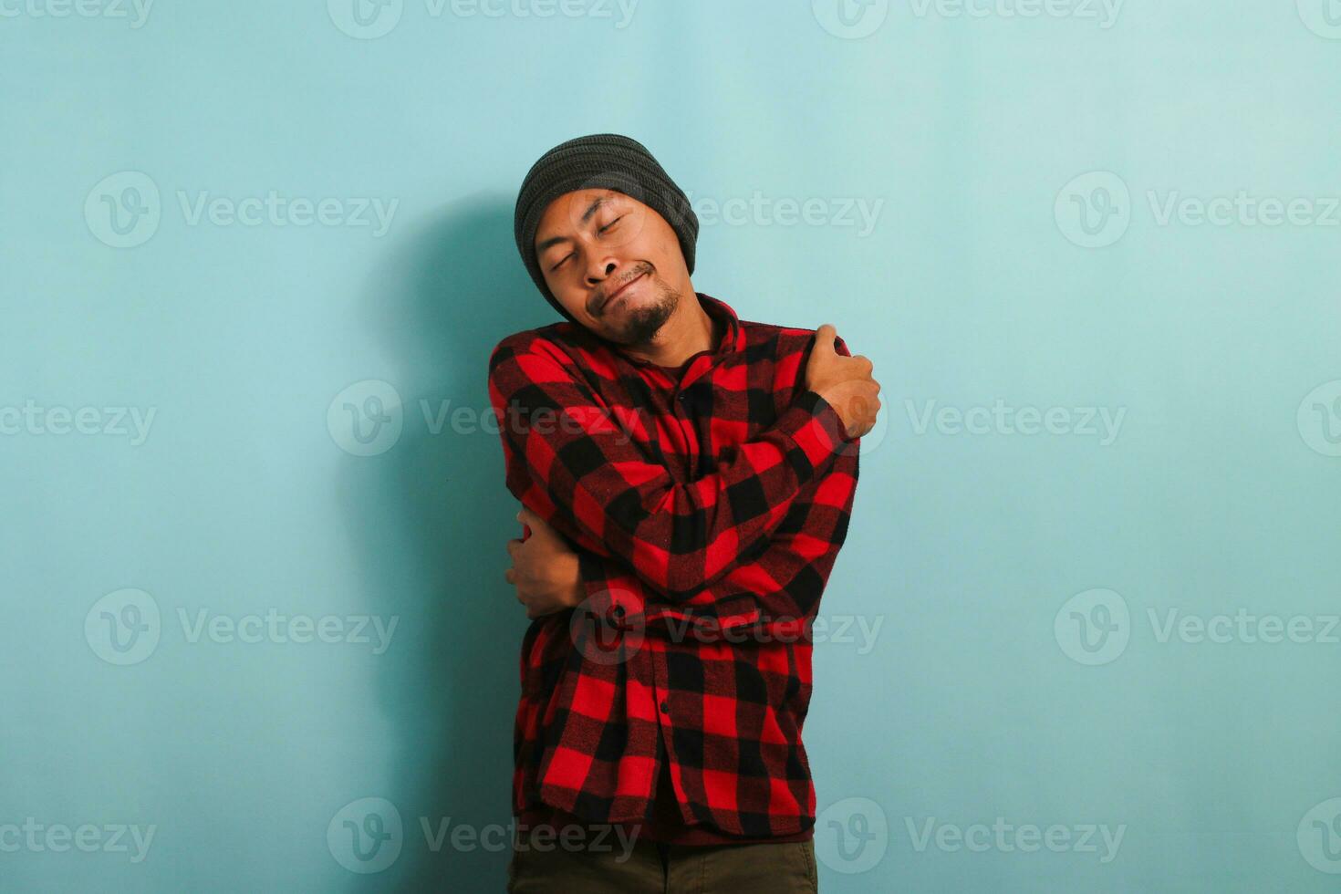 Young Asian man with beanie hat and red plaid flannel shirt hugs himself, expressing happiness and positivity, while standing against a blue background. Self love and self care concept. photo