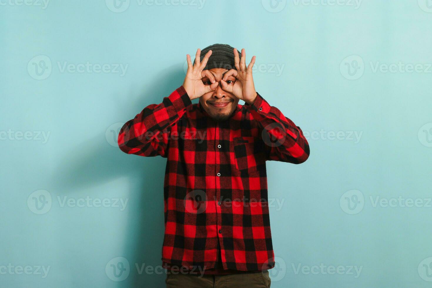 emocionado joven asiático hombre con gorro sombrero y rojo tartán franela camisa hace el Okay firmar gesto con su dedos, interino como prismáticos con su ojos echar un vistazo mediante a ellos, aislado en azul antecedentes foto