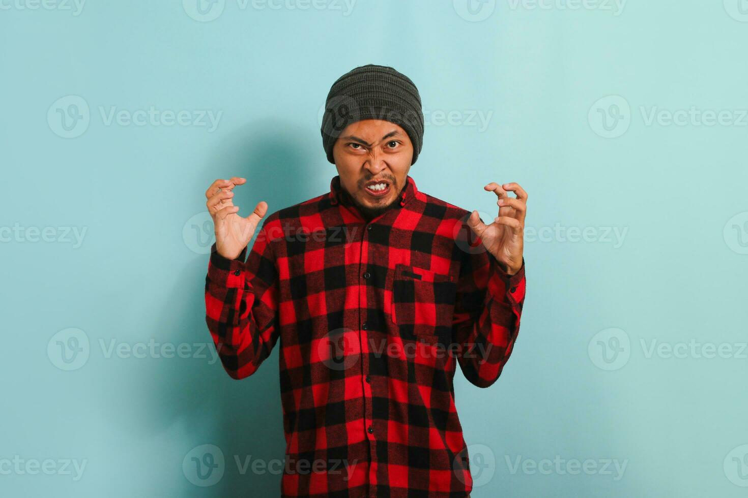 An angry young Asian man with a beanie hat and a red plaid flannel shirt holds his hands in a furious gesture, screaming at the camera. He is isolated on a blue background photo