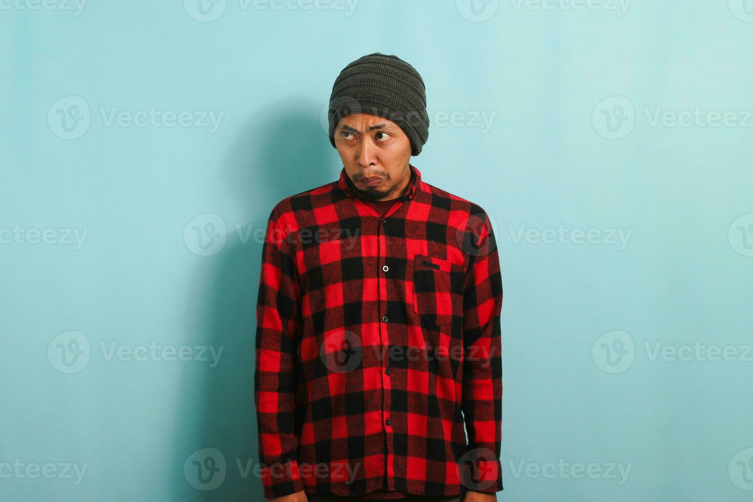 Displeased young Asian man with a beanie hat and a red plaid flannel shirt looks aside at an empty copyspace with a disgusted expression while standing against a blue background photo