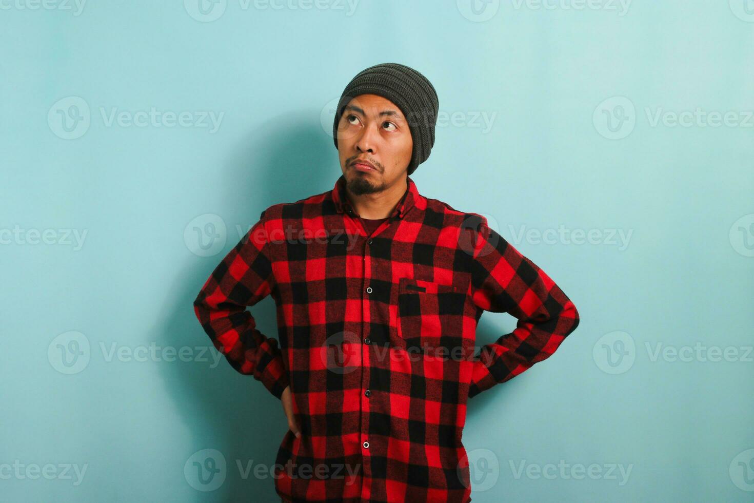 A bored young Asian man wearing a beanie hat and a red plaid flannel shirt stands with an arm akimbo gesture, looking to the left deep in thought about what to do, isolated on a blue background photo