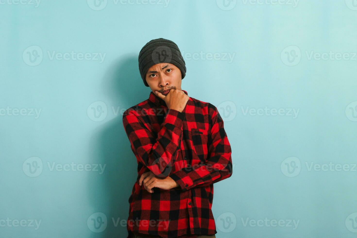 A pensive young Asian man with a beanie hat and a red plaid flannel shirt keeps his hand on his chin, looking serious and worried as he gazes at the camera while standing against a blue background photo