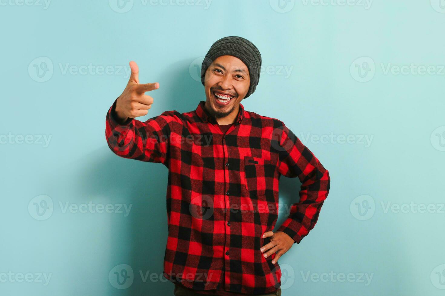 confidente joven asiático hombre con un gorro sombrero y un rojo tartán franela camisa es señalando su dedo a el cámara y sonriente, eligiendo, atractivo gente, o reclutamiento. él es aislado en azul antecedentes foto