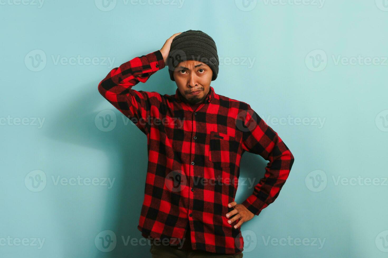 An unhappy young Asian man with a beanie hat and a red plaid flannel shirt grabs his head, staring alarmed at the camera, and feeling anxious while standing against a blue background photo