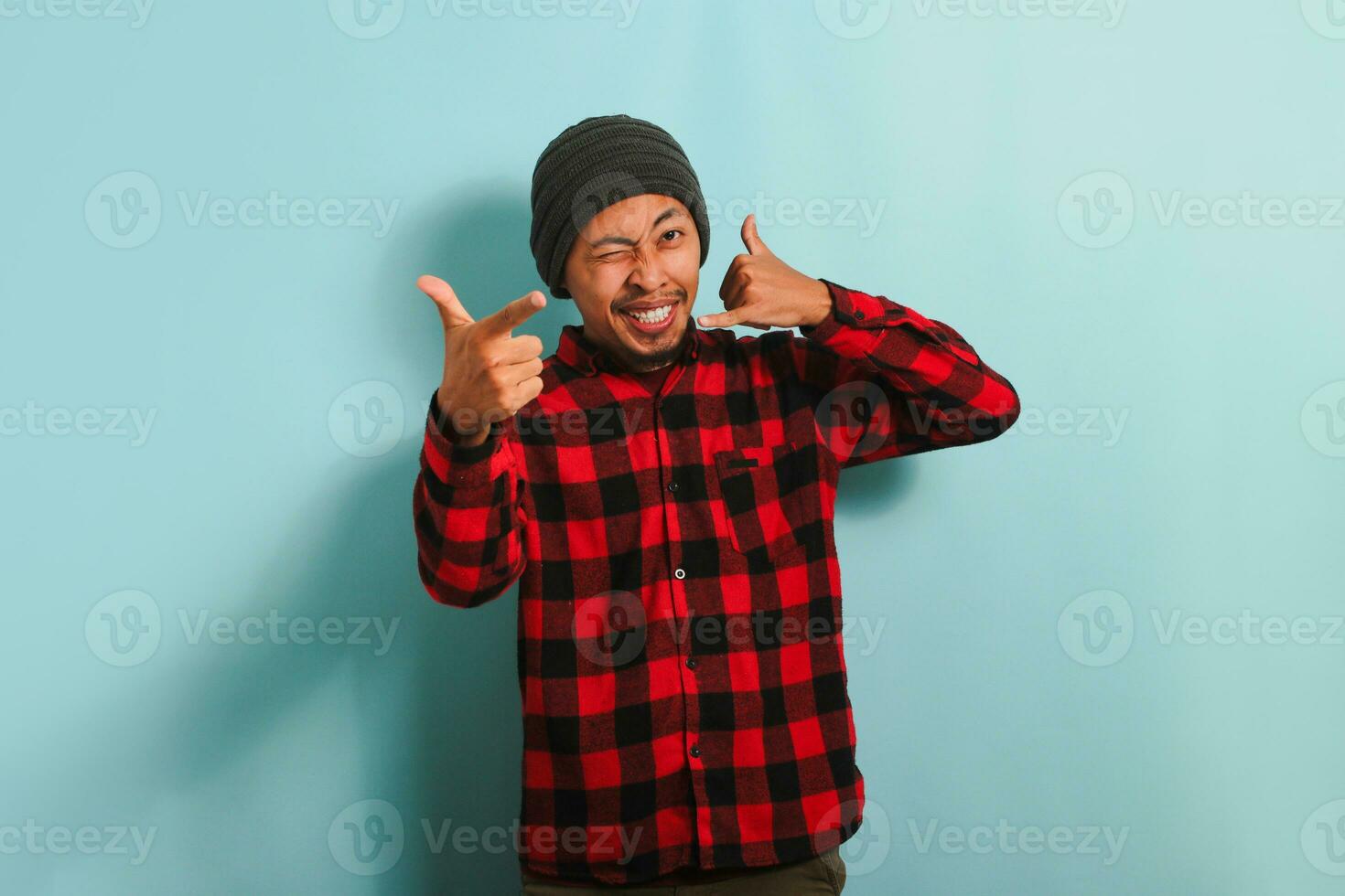 An excited young Asian man wearing a beanie hat and a red plaid flannel shirt makes a call me gesture while blinking and pointing at the camera, isolated on a blue background photo