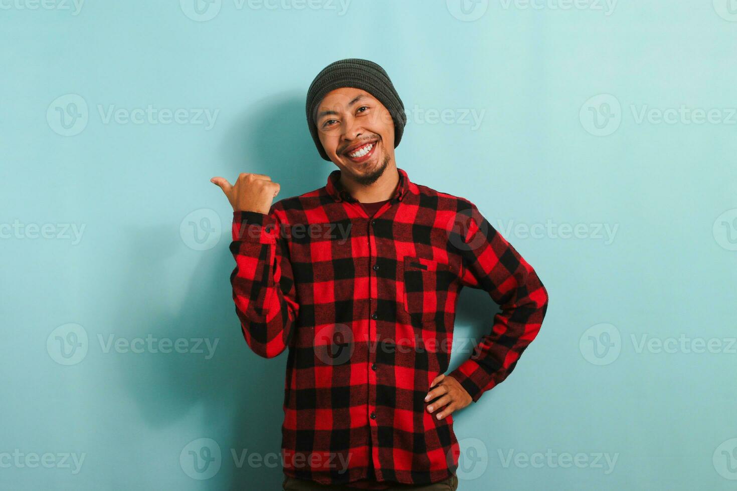 Excited Young Asian man with a beanie hat and red plaid flannel shirt is pointing aside with his thumb, indicating the copy space, isolated on a blue background photo